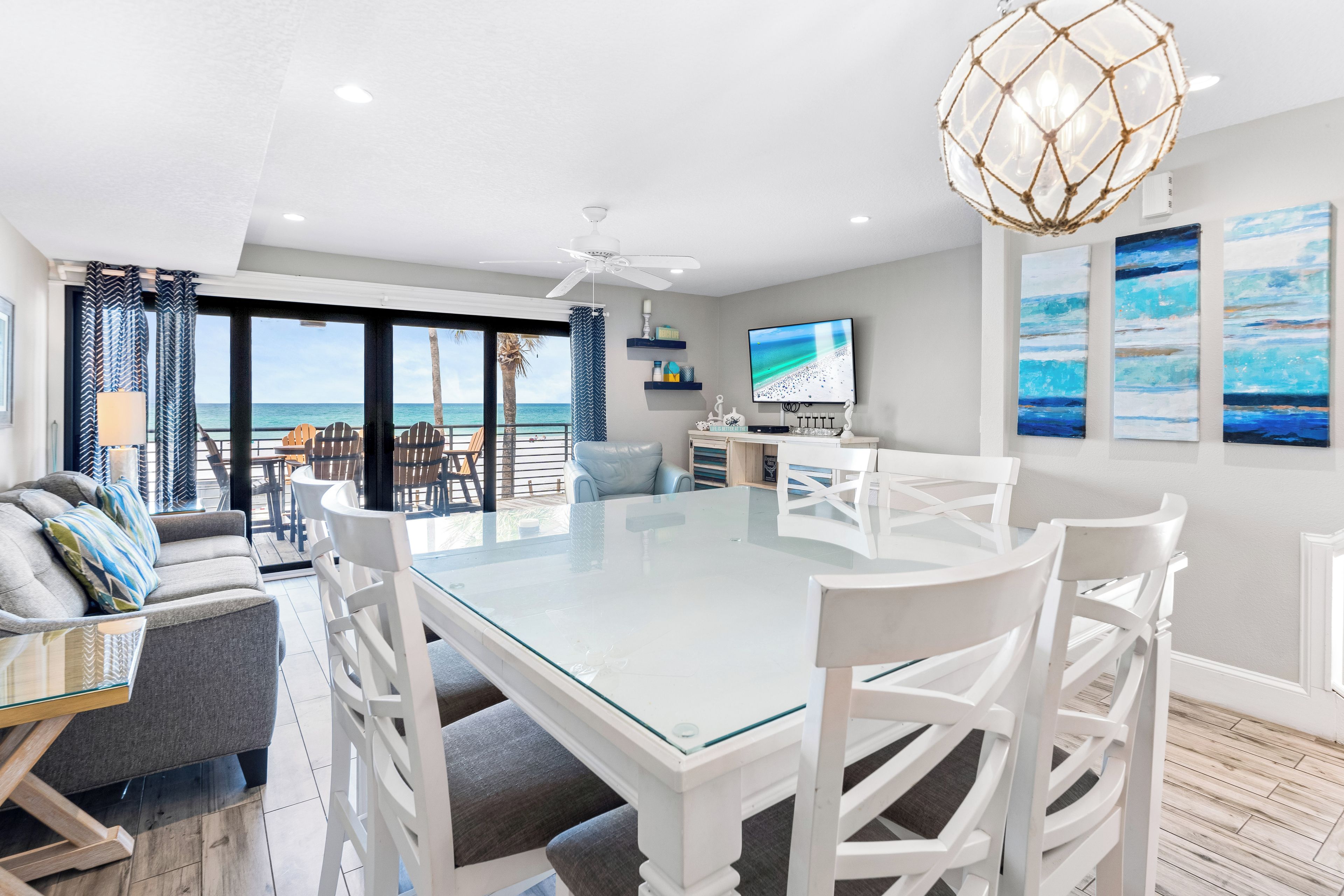 A spacious living area with a glass-top table, white chairs, a ceiling fan, and a TV.