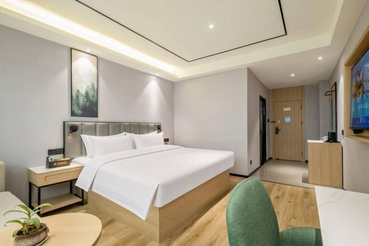 A modern hotel room with a large bed, bedside tables, a green chair, and a TV.