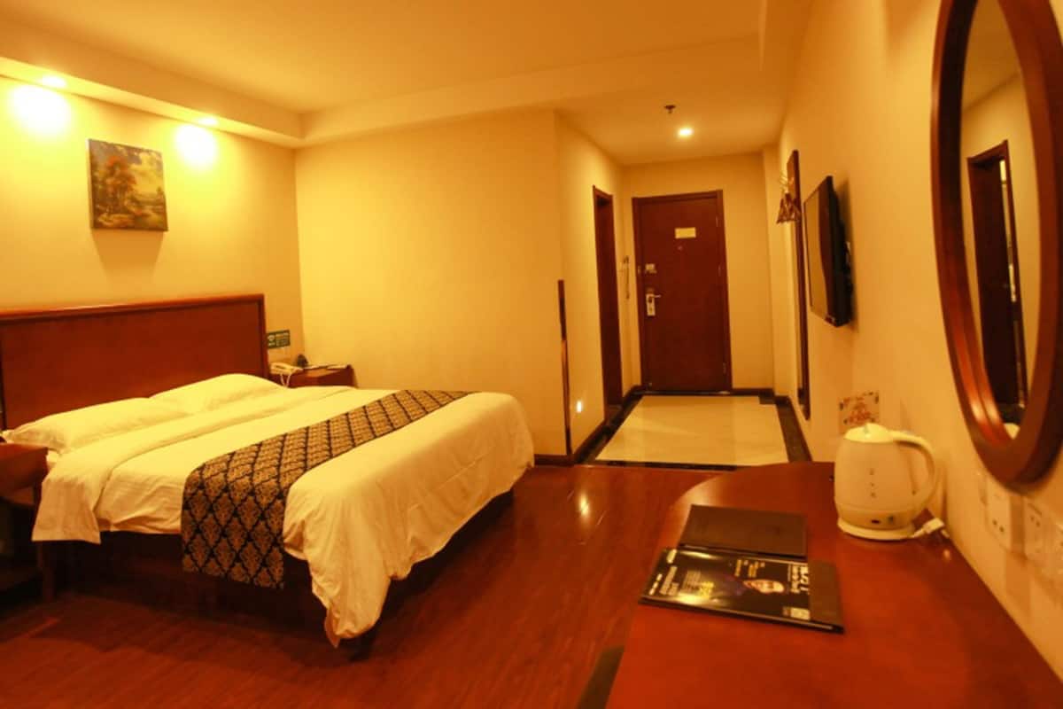 A hotel room with a double bed, a bedside table with a lamp, a desk with a computer, a mirror, and a door leading to another room.