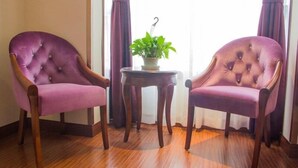 Deux fauteuils violets à dossier capitonné et à pieds en bois, une petite table en bois sur laquelle est posée une plante en pot, et une fenêtre ornée de rideaux violets.