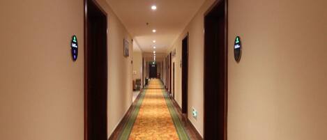 A hotel hallway with a carpeted floor, doors on both sides, and illuminated signs indicating room numbers.
