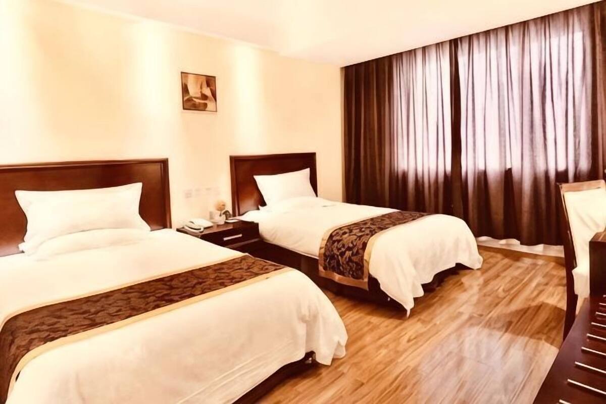 Habitación de hotel con dos camas, suelo de madera, una mesita de noche con lámpara y un ventanal grande con cortinas.