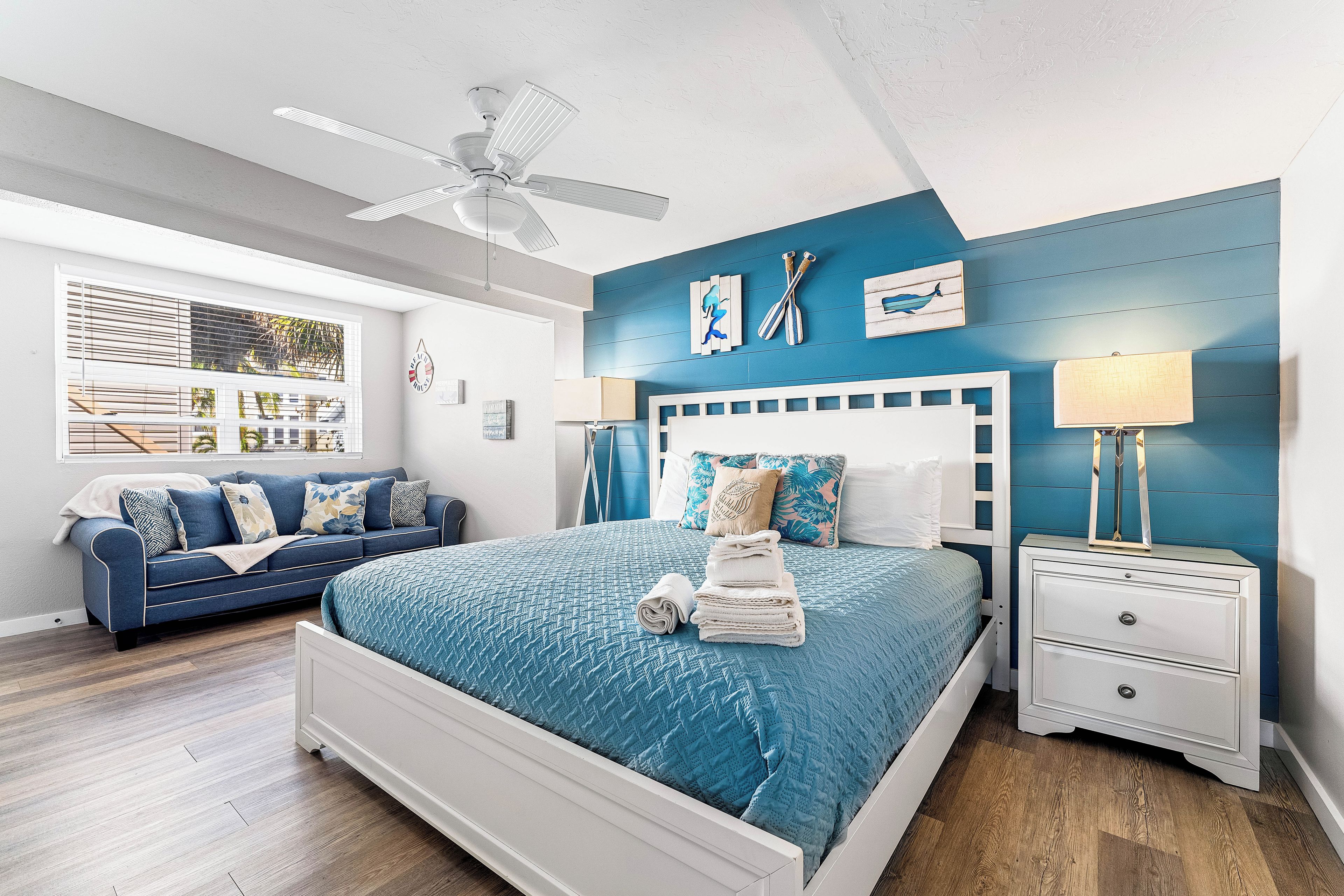 A bedroom with a bed, a nightstand, a ceiling fan, a window with blinds, and a blue wall.