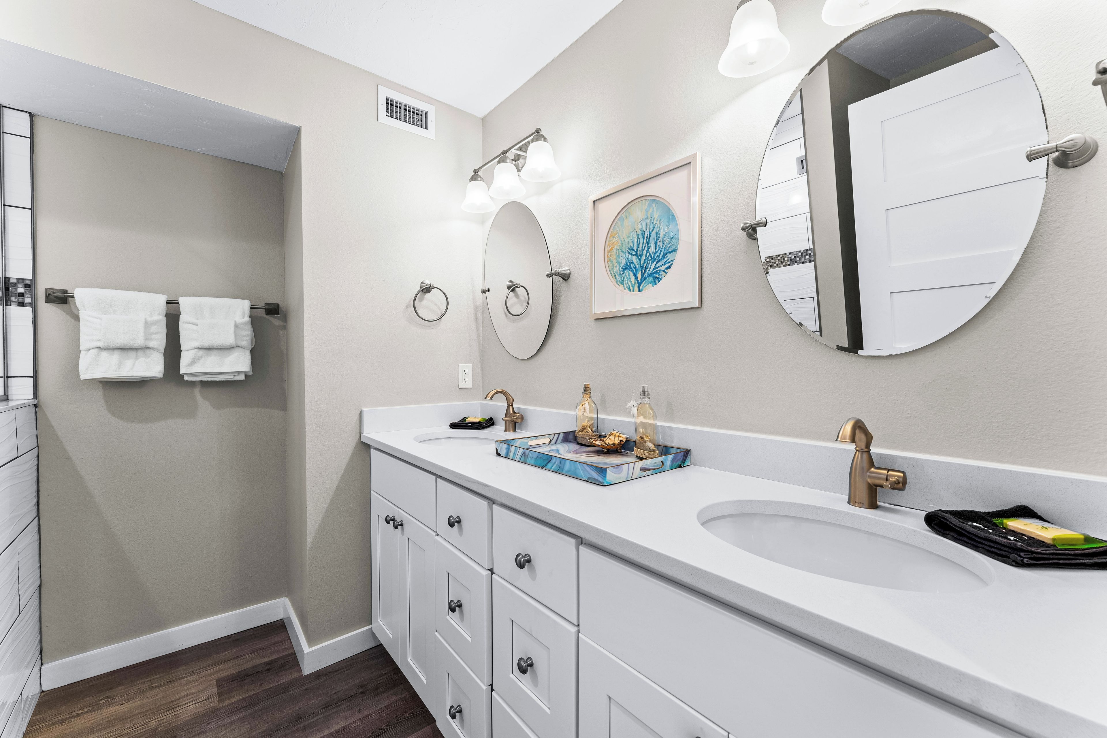 A modern bathroom with a large white sink, two mirrors, a framed artwork, and a towel rack with two towels.
