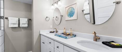 A modern bathroom with a large white sink, two mirrors, a framed artwork, and a towel rack with two towels.