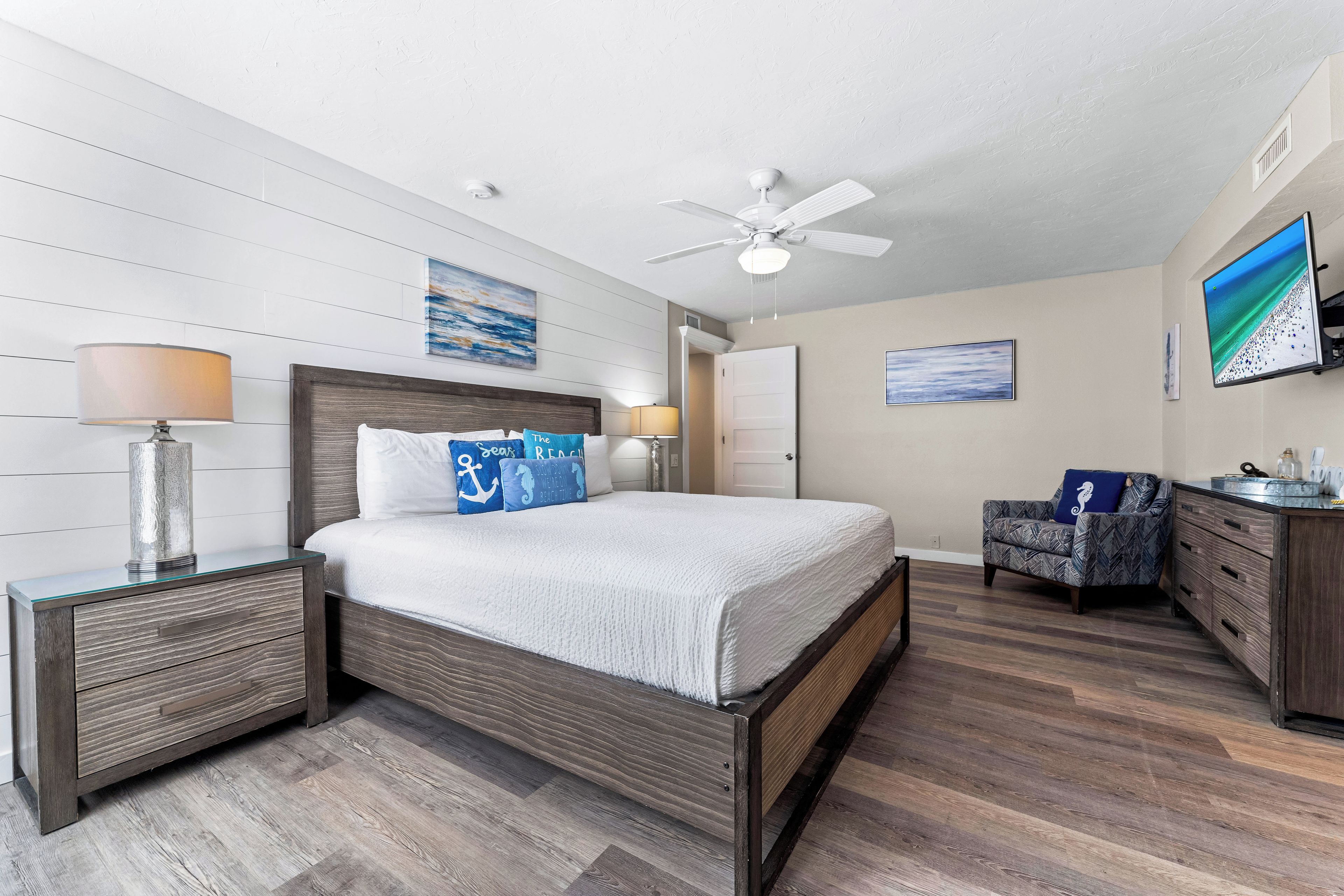 A bedroom with a bed, nightstands, a desk, a chair, a ceiling fan, and artwork on the walls.