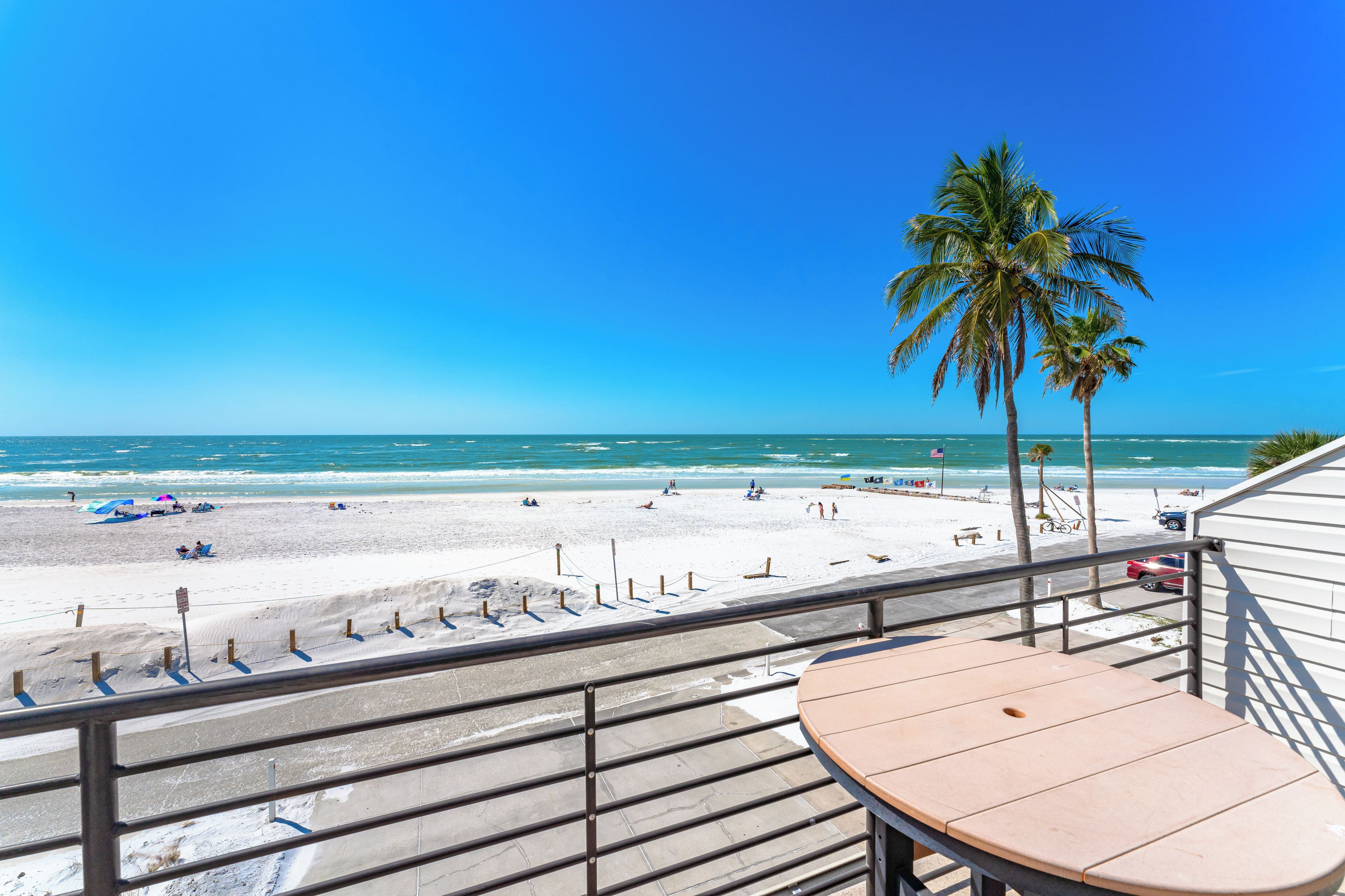 Une vue sur la plage depuis un balcon, avec une table et des chaises, donnant sur une plage de sable bordée de palmiers et sur l’océan.