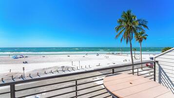 Une vue sur la plage depuis un balcon, avec une table et des chaises, donnant sur une plage de sable bordée de palmiers et sur l’océan.