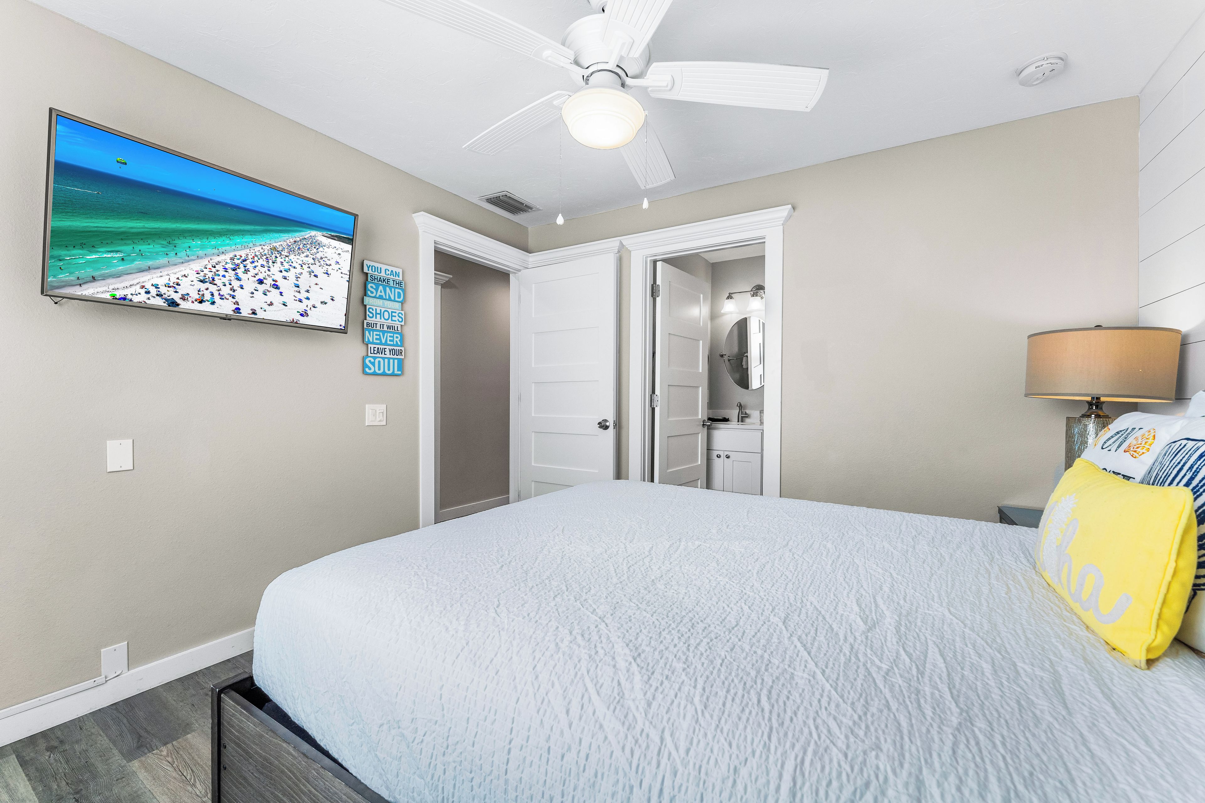 Un dormitorio con cama, ventilador de techo, televisión en la pared y baño visible a través de una puerta abierta.