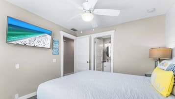 Un dormitorio con cama, ventilador de techo, televisión en la pared y baño visible a través de una puerta abierta.