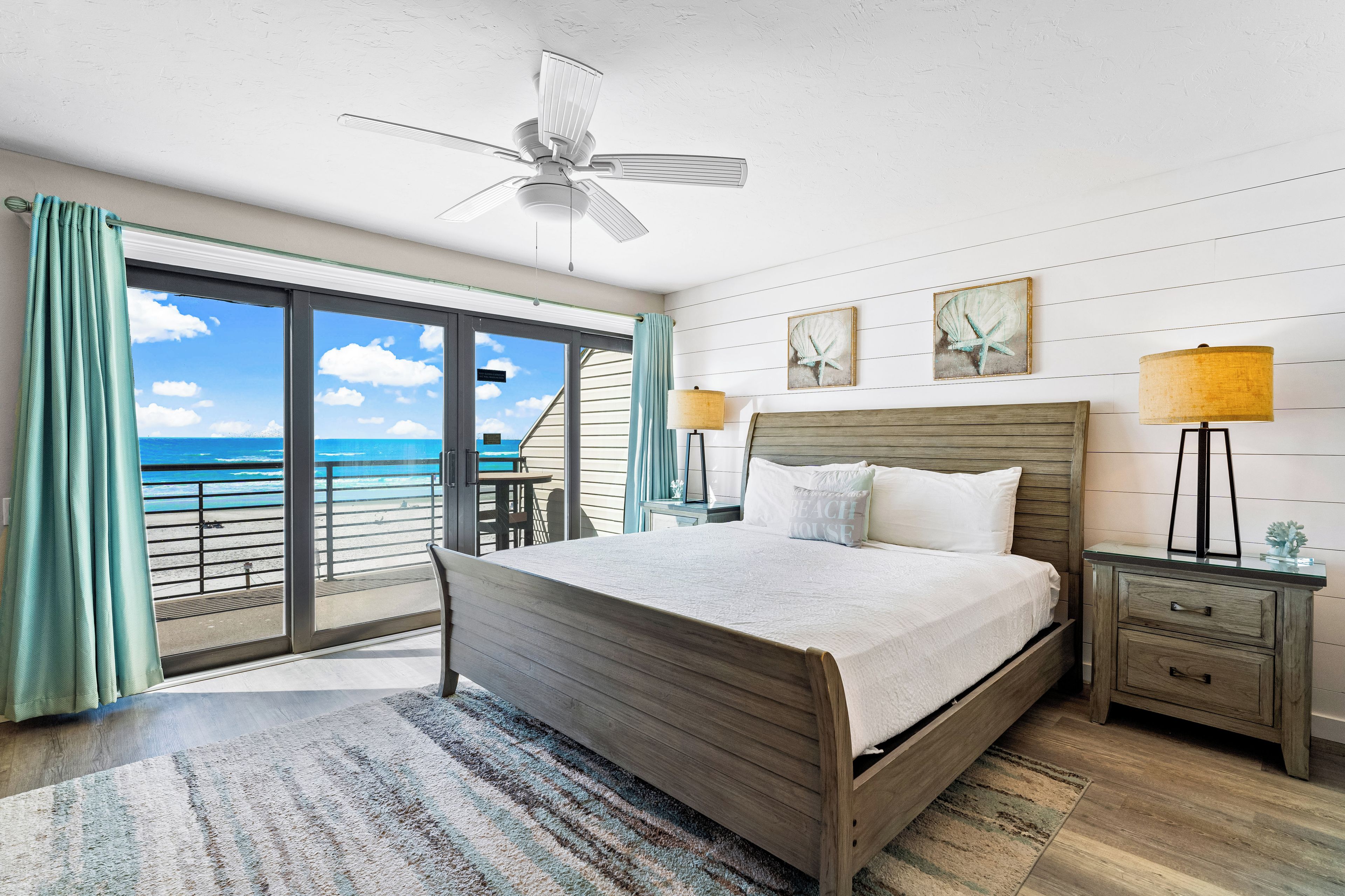 Une chambre à coucher avec un lit, une table de chevet, une lampe, un ventilateur de plafond et une vue sur la plage, visible à travers de grandes fenêtres.