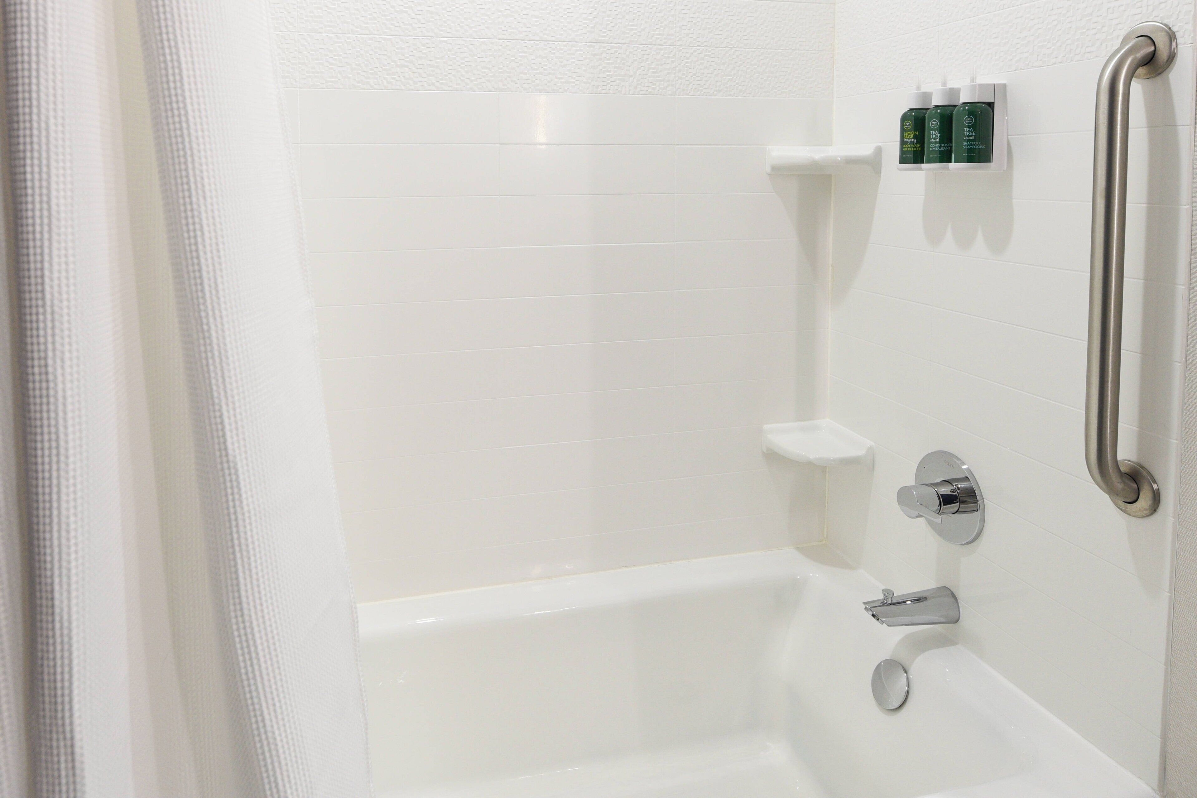 A white bathroom with a bathtub, a handrail, and a shelf with toiletries.