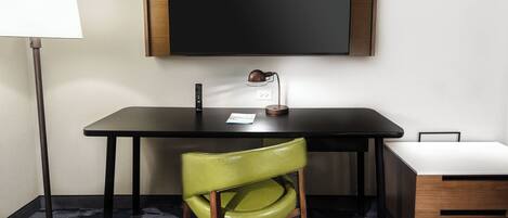 A modern hotel room with a desk, a green chair, a lamp, and a TV mounted on the wall.