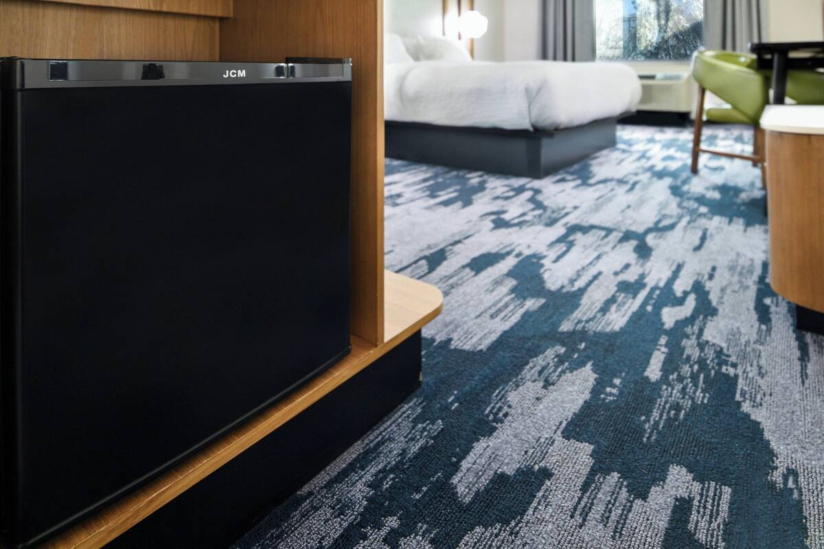 A hotel room with a JCM refrigerator, a bed with white bedding, a green chair, and a blue patterned carpet.
