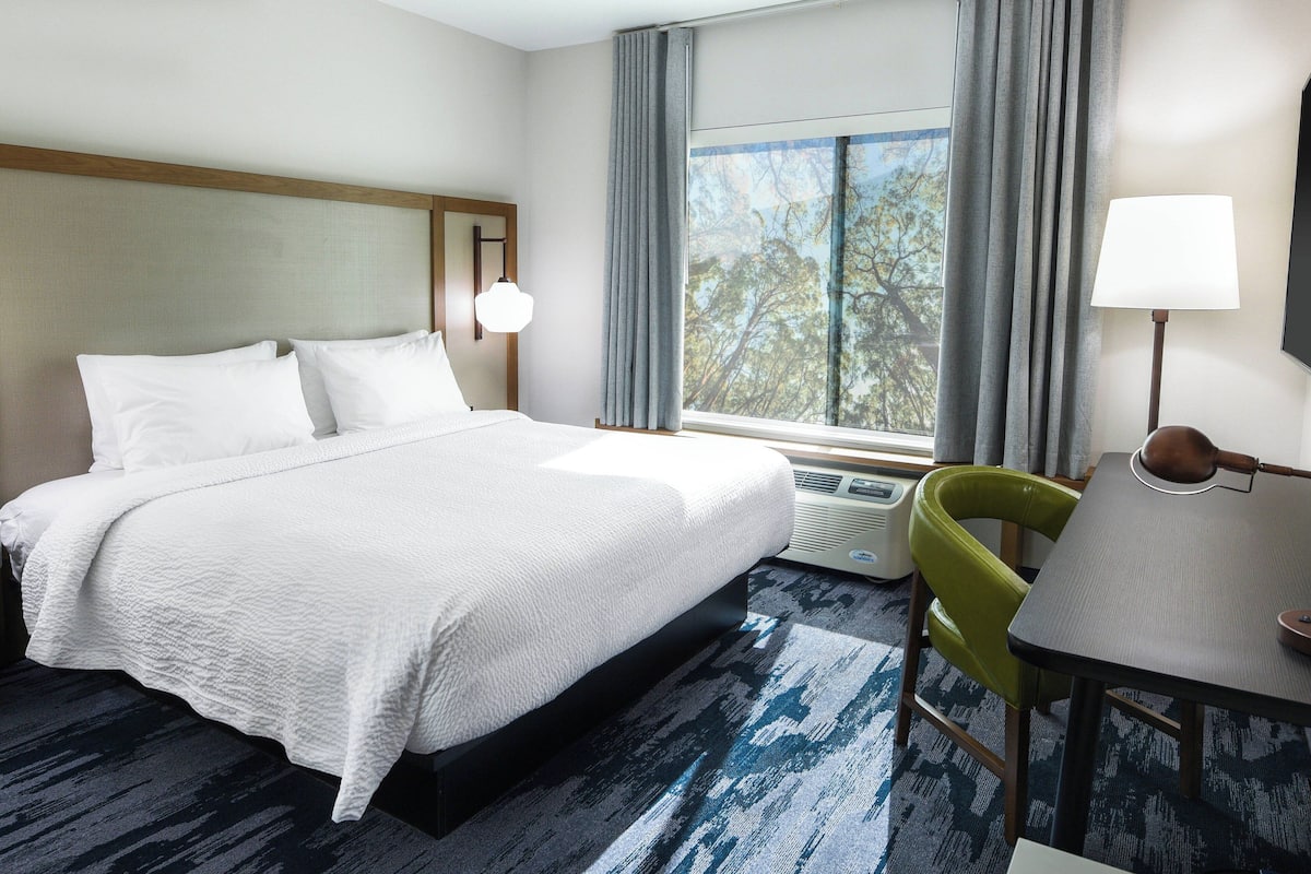 A hotel room with a bed, a desk, a chair, a lamp, and a window with curtains.