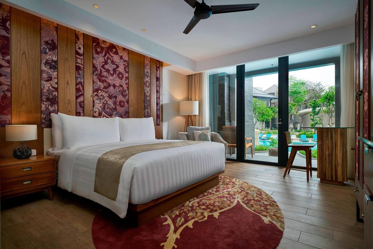 Une chambre spacieuse avec un grand lit, des boiseries, un ventilateur de plafond et une vue sur une piscine et un espace de détente extérieur.