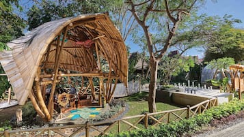 A bamboo structure with a curved roof, surrounded by a garden with a chessboard, trees, and a fence.