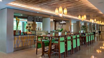 A modern cafe with wooden ceiling panels, green upholstered chairs, and pendant lighting.