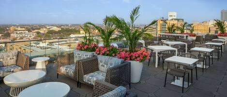 A rooftop terrace with wicker furniture, white tables, and potted plants.