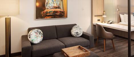 A modern hotel room with a grey sofa, a wooden side table, a desk with a chair, a large framed artwork of a horse, and a lamp.