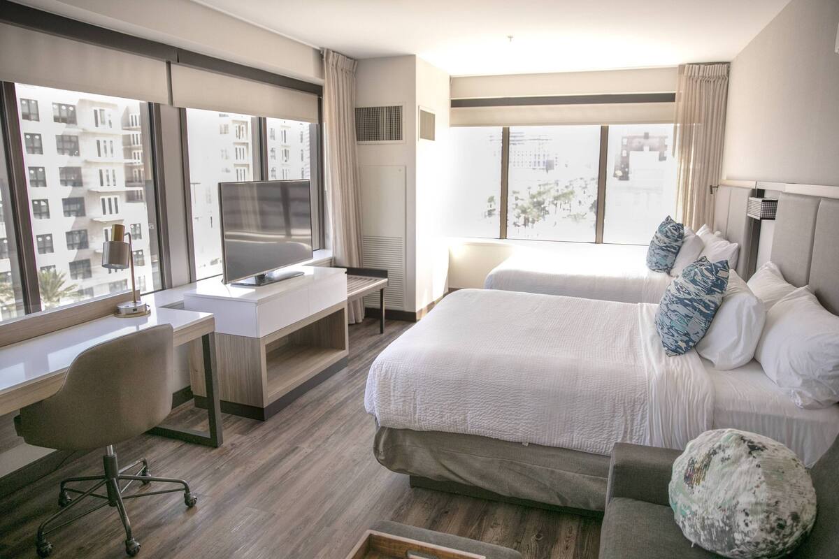A hotel room with a large bed, a desk with a computer, a TV, and a view of the city through large windows.