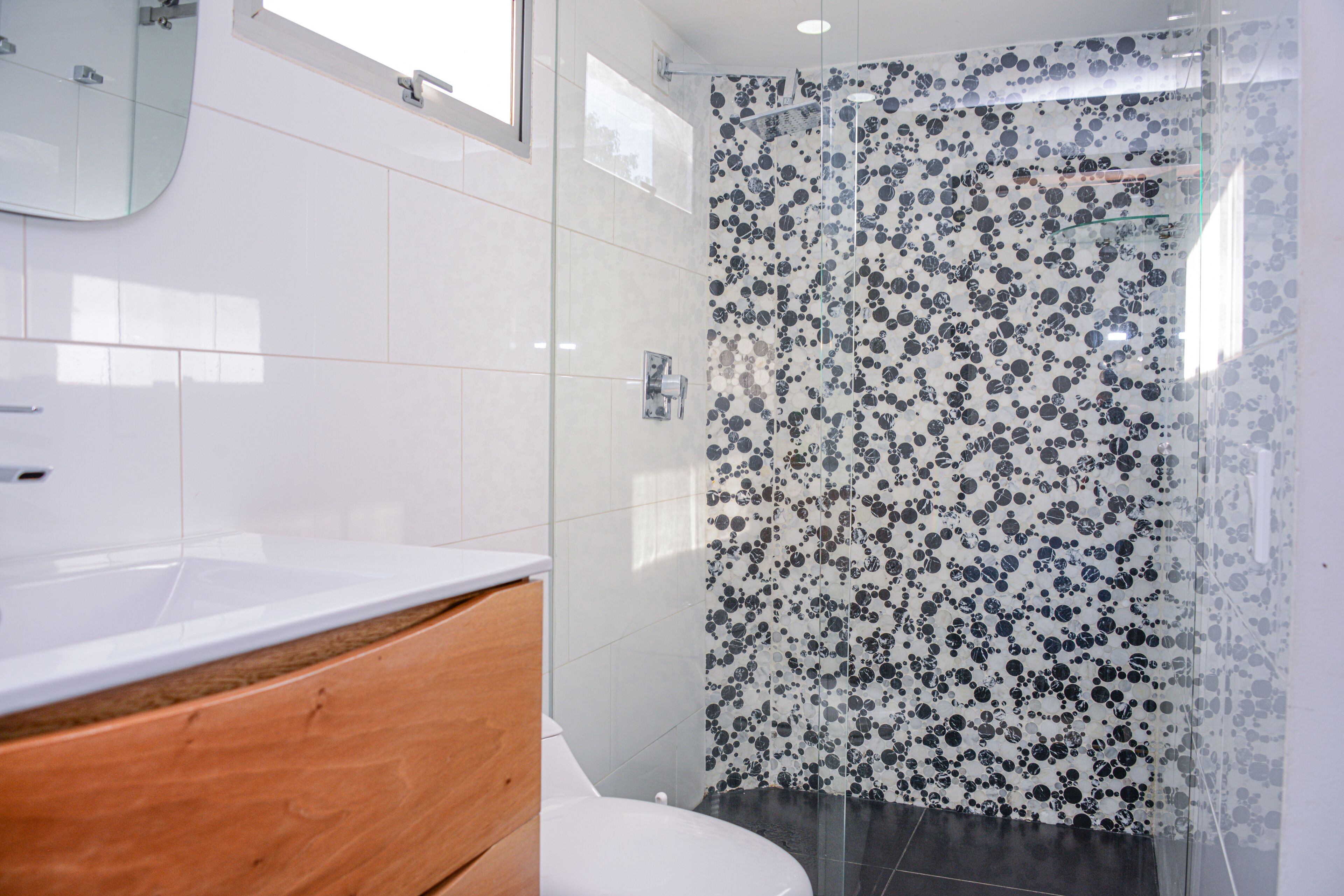 Un baño moderno con lavamanos blanco, tocador de madera y ducha con puerta de vidrio estampada en blanco y negro.