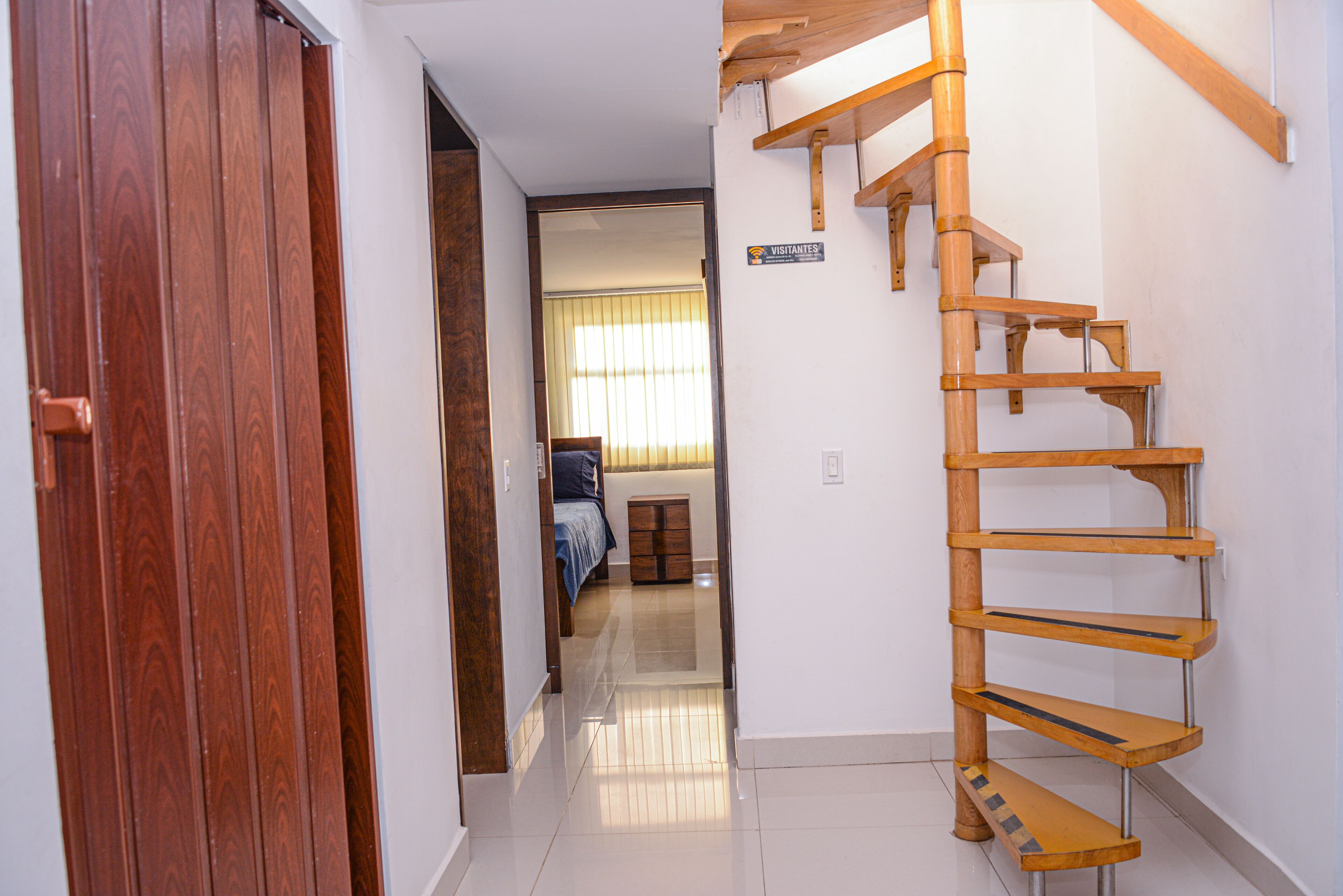 A wooden spiral staircase in a hallway with a wooden door on the left and a sign reading "VISITANTES" on the wall.