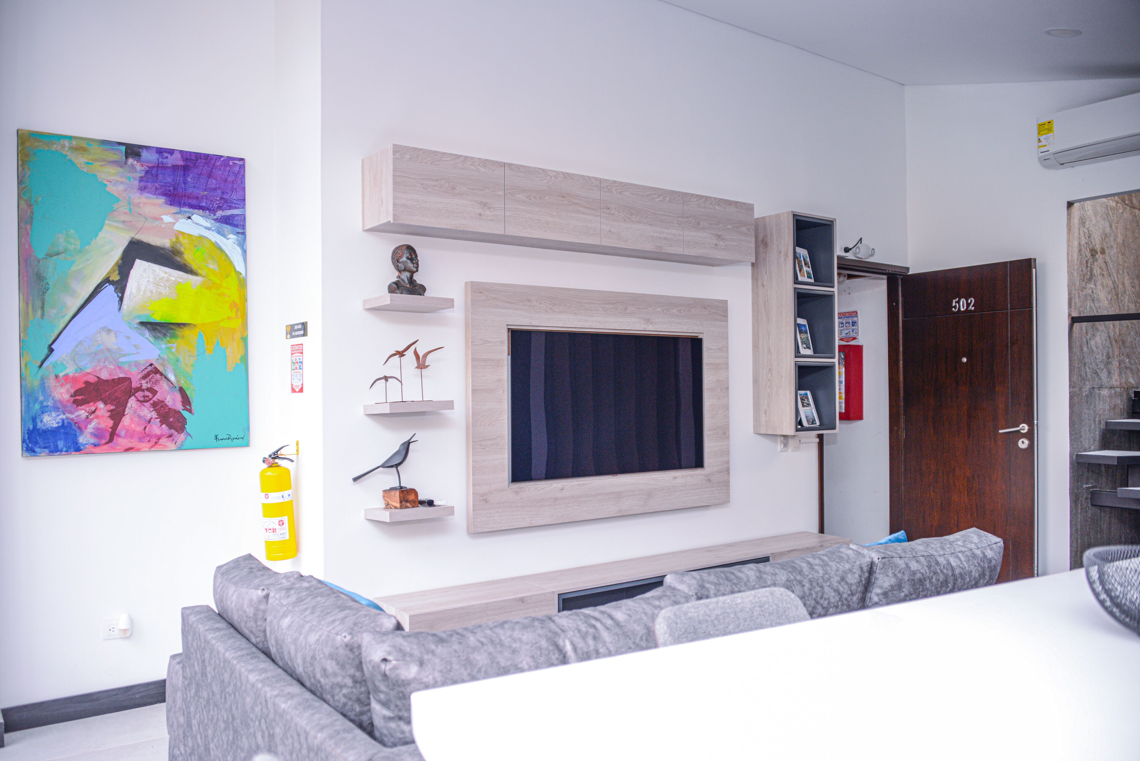 Una sala moderna con un televisor grande, sofás grises, un cuadro abstracto de colores y una puerta de madera con el número 502.