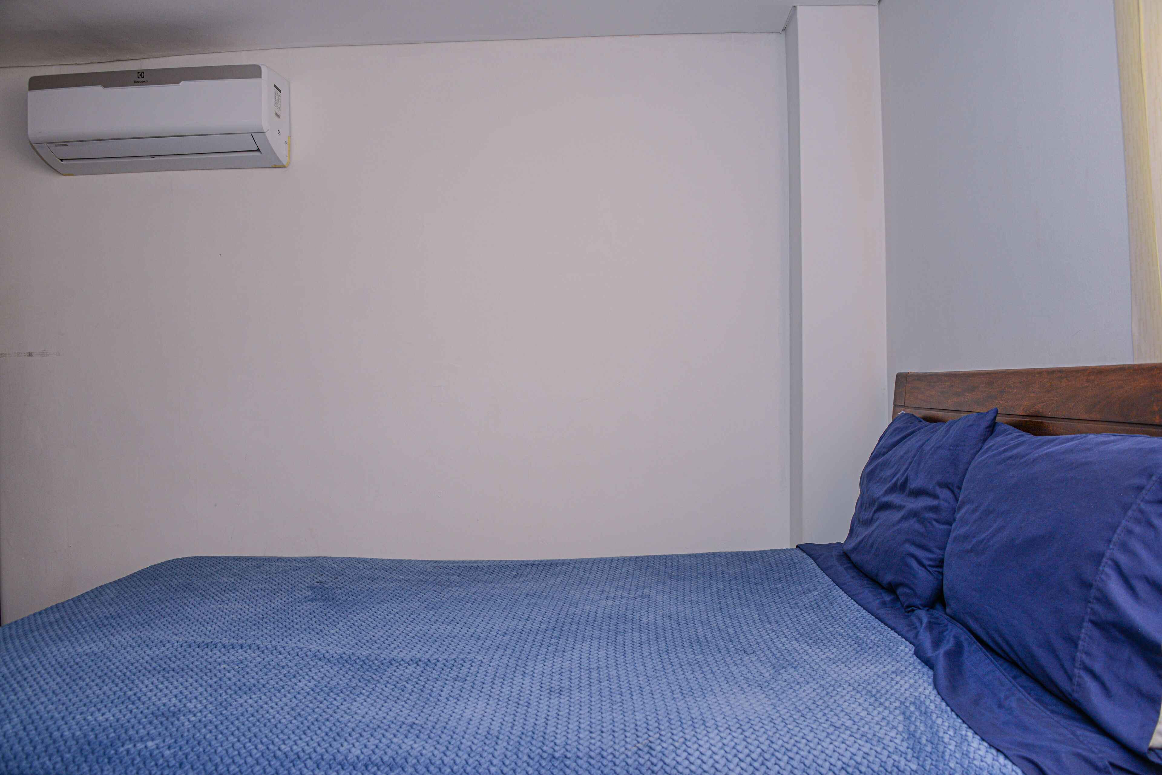 En enkeltseng med blå madras og puder, et træhovedgærde, og en hvid væg med en hvid aircondition-enhed monteret på den.