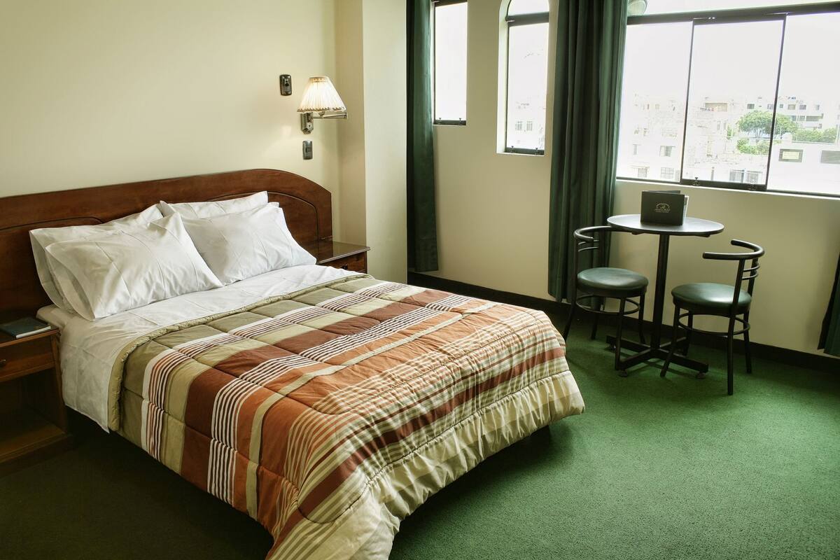 A hotel room with a bed, nightstand, lamp, two chairs, a small table, and a window with curtains.