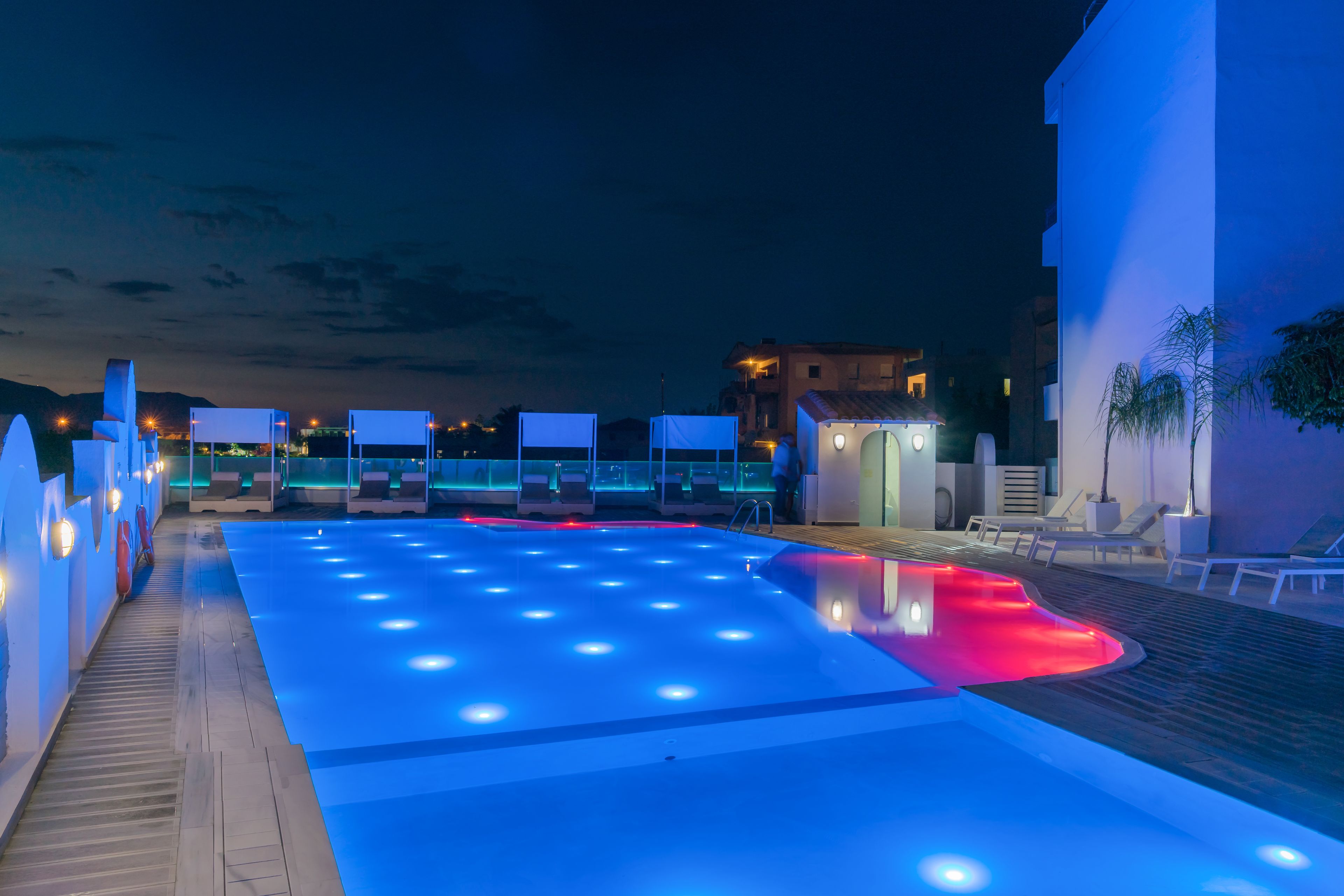 Une piscine illuminée de lumières bleues et rouges, entourée de chaises longues et d’une terrasse en bois.
