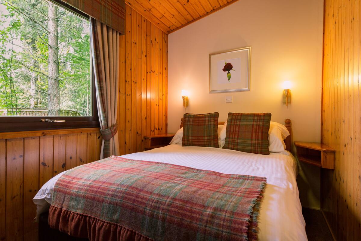 A wooden-paneled bedroom with a bed, plaid bedding, a framed picture on the wall, and a window with curtains.