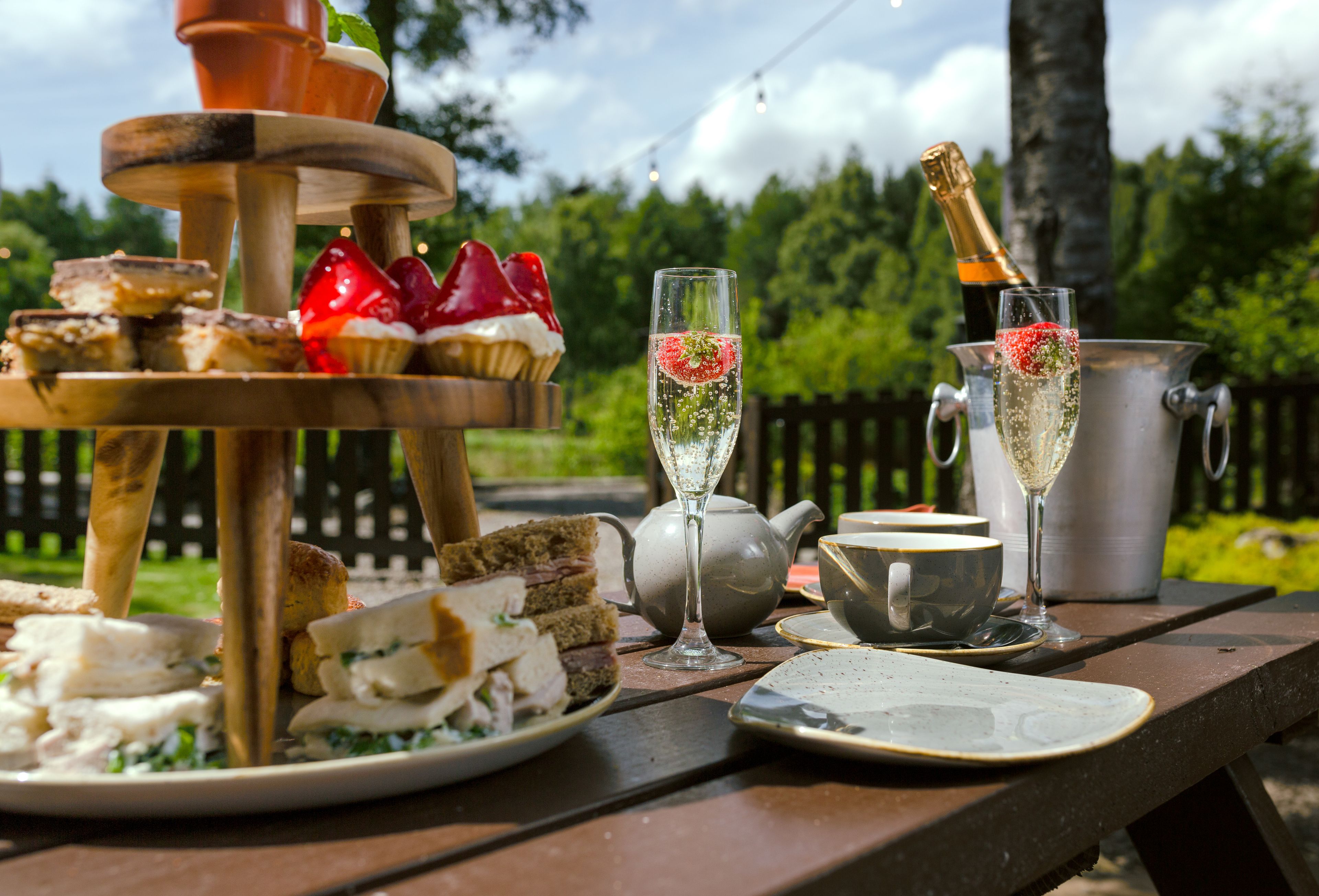 Un'apparecchiatura da picnic con un vassoio a più livelli per dolci, calici da champagne e un teiera su un tavolo di legno.