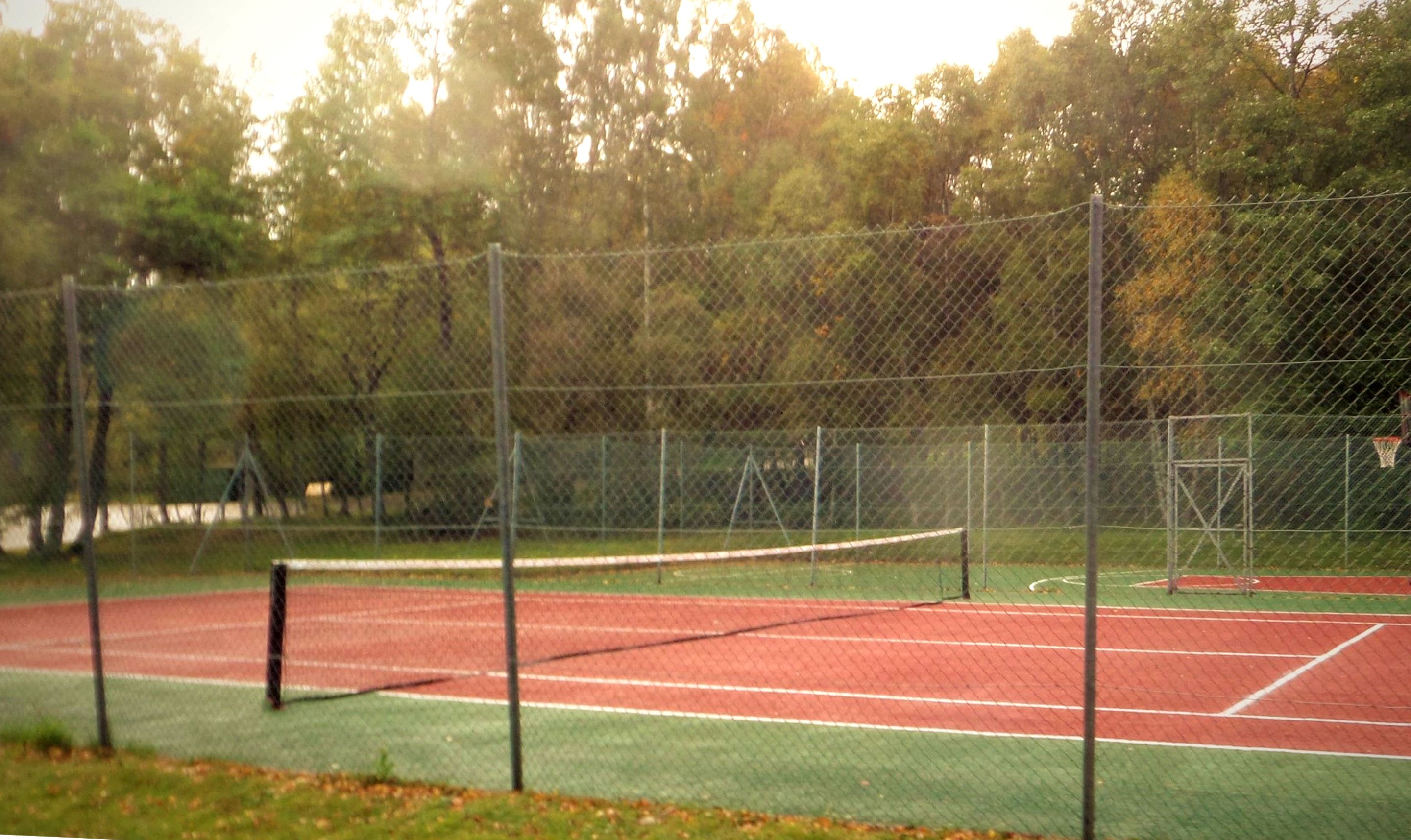 A tennis court with a net, surrounded by a chain-link fence and trees.