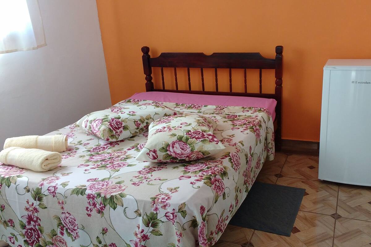 Una cama con ropa de cama floral, dos toallas enrolladas, un cabecero de madera y un refrigerador blanco en una habitación con paredes anaranjadas y suelo de madera.