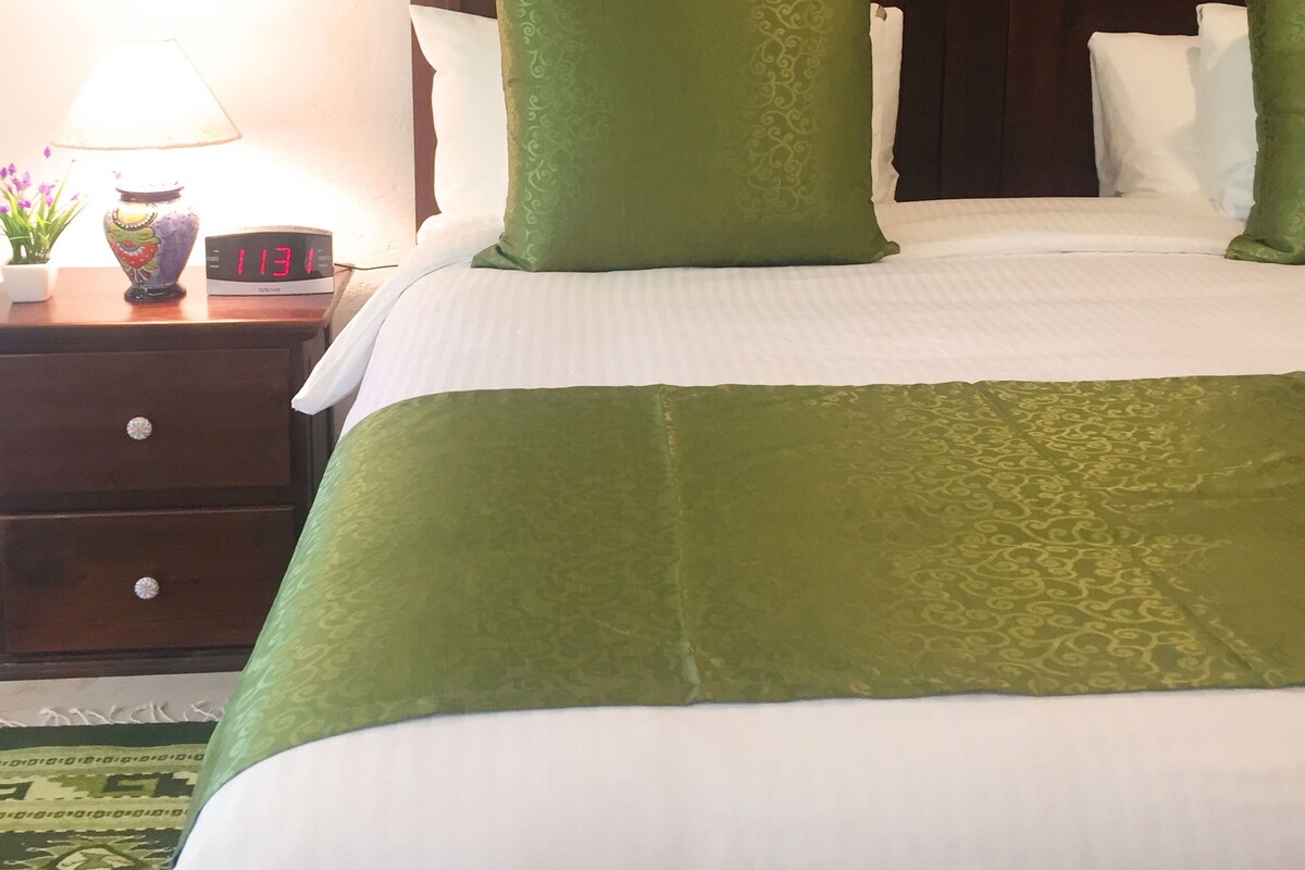 Una cama bien hecha con ropa de cama verde y blanca, un cabecero de madera, una mesita de noche con una lámpara y un reloj digital, y un pequeño jarrón con flores.