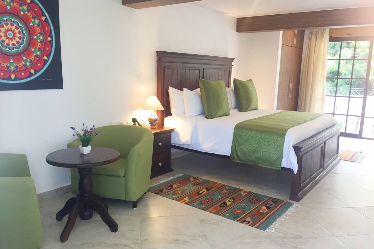 A bedroom with a bed, green armchair, bedside table with lamp, and a colorful rug.
