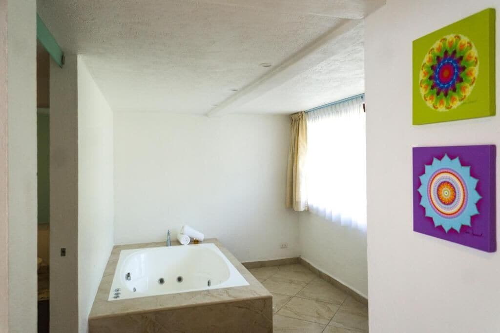 A bathroom with a large bathtub, a window with curtains, and two colorful wall art pieces.