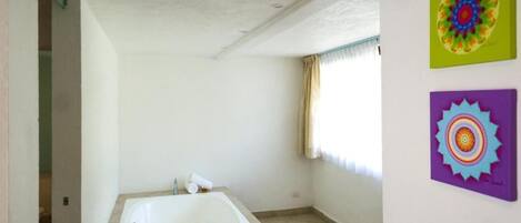 A bathroom with a large bathtub, a window with curtains, and two colorful wall art pieces.