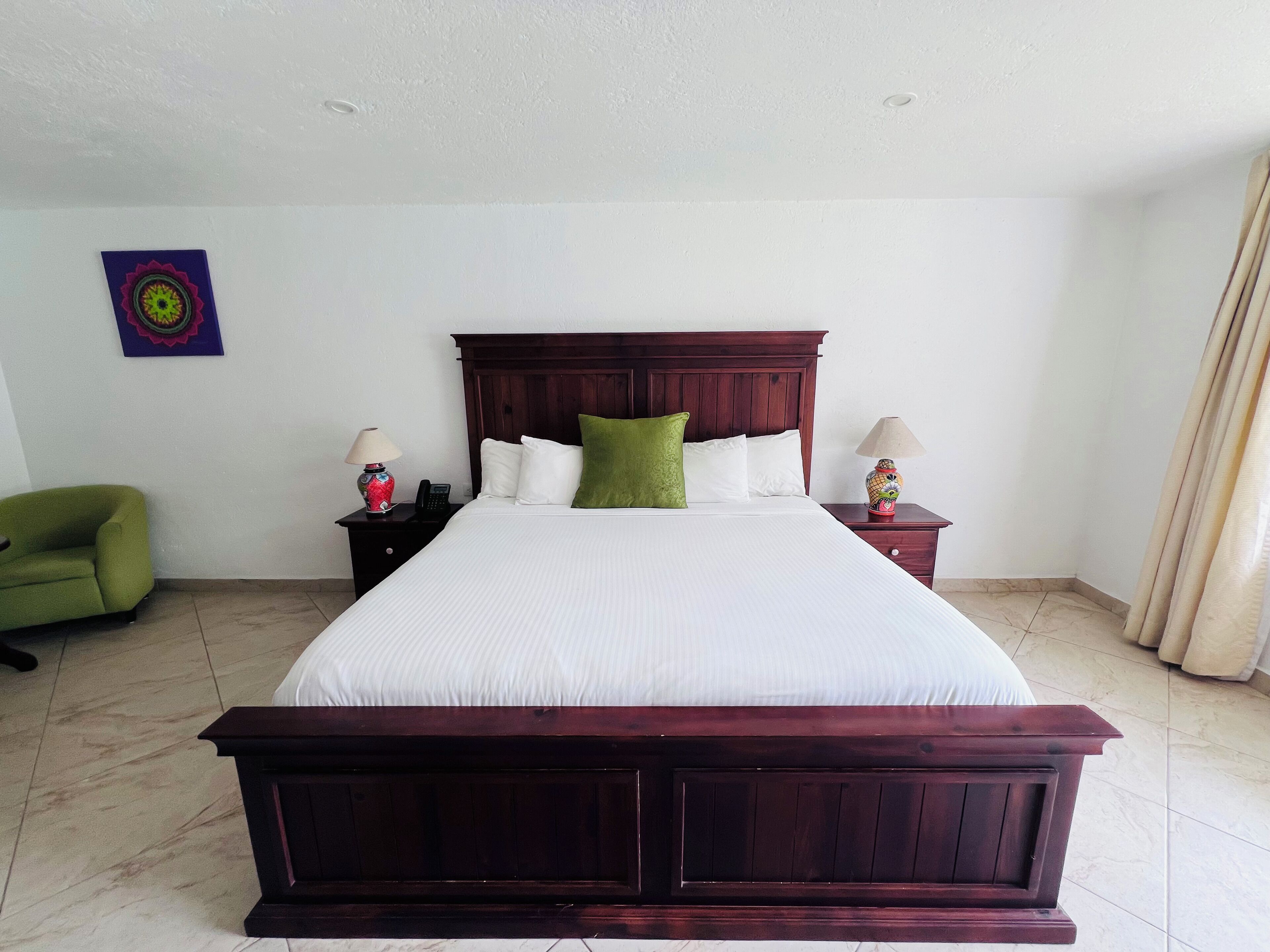 A neatly made bed with a wooden headboard, two bedside tables with lamps, a green chair, and a decorative wall piece.