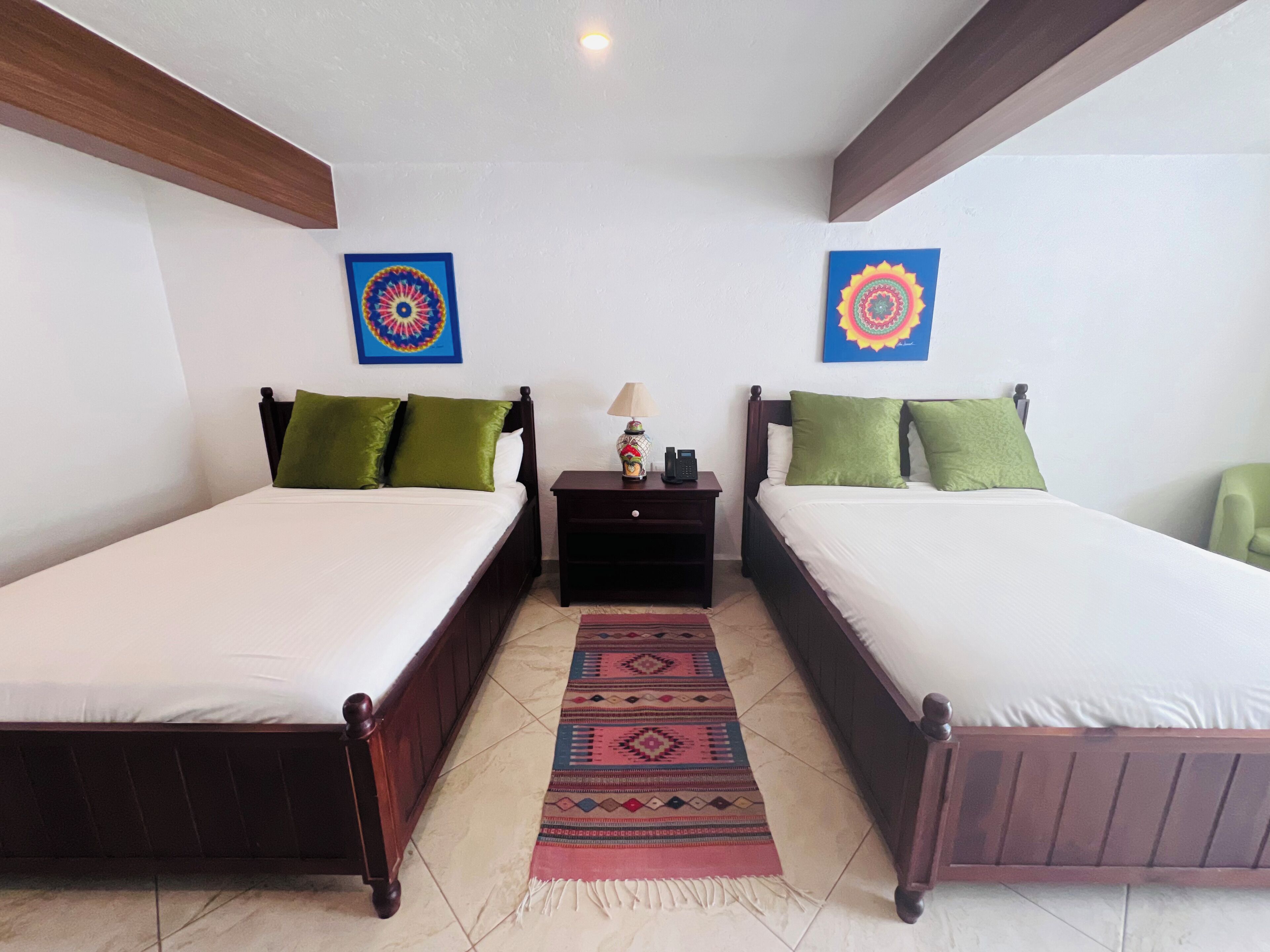A double room with two beds, each with green pillows, a nightstand with a lamp, and a patterned rug.