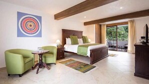 Habitación de hotel con cama, dos sillones verdes, una mesita, alfombra colorida y un ventanal con vistas a la vegetación.