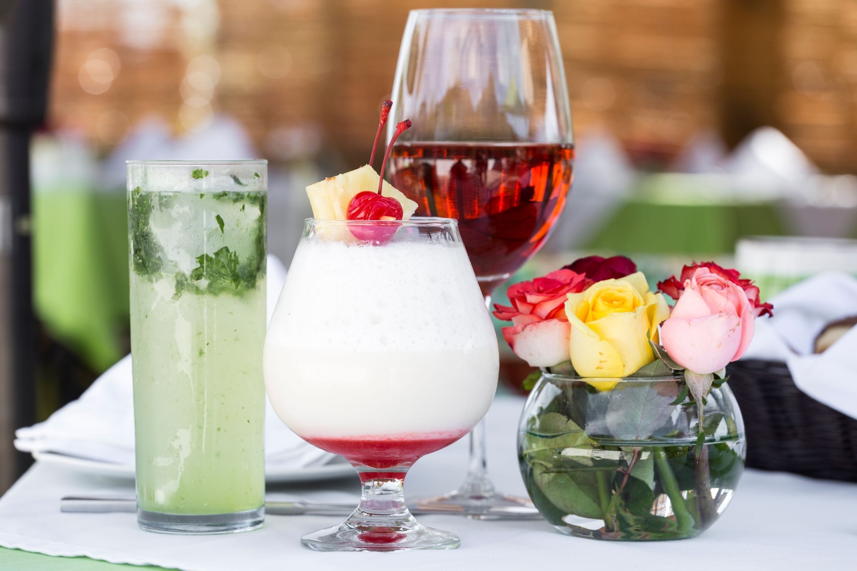 Three glasses of beverages, a wine glass, and a vase with flowers on a table.
