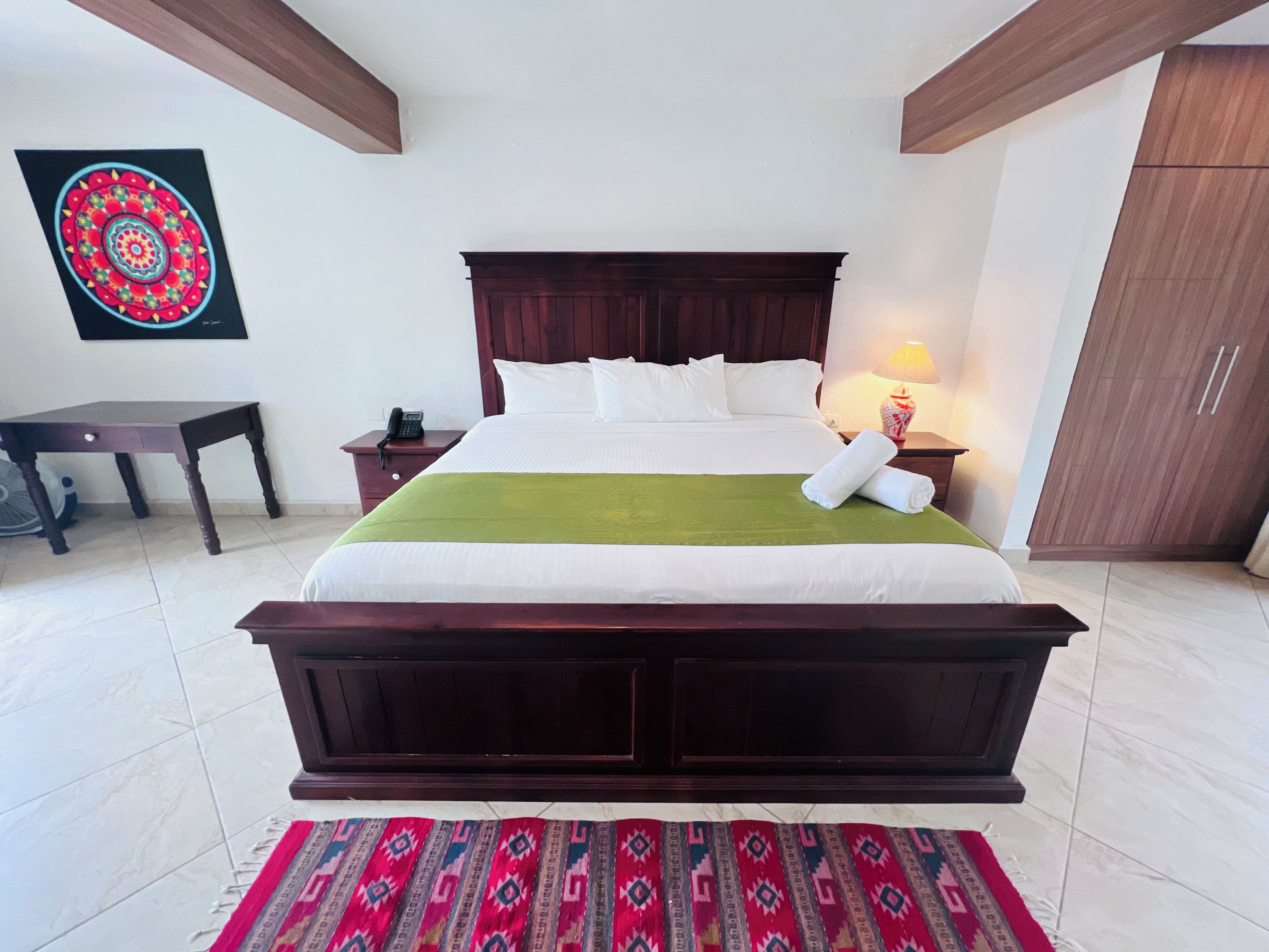 A neatly made bed with white linens, a green throw blanket, and a rolled towel on the side. There is a wooden nightstand with a lamp and a phone. A patterned rug is on the floor.
