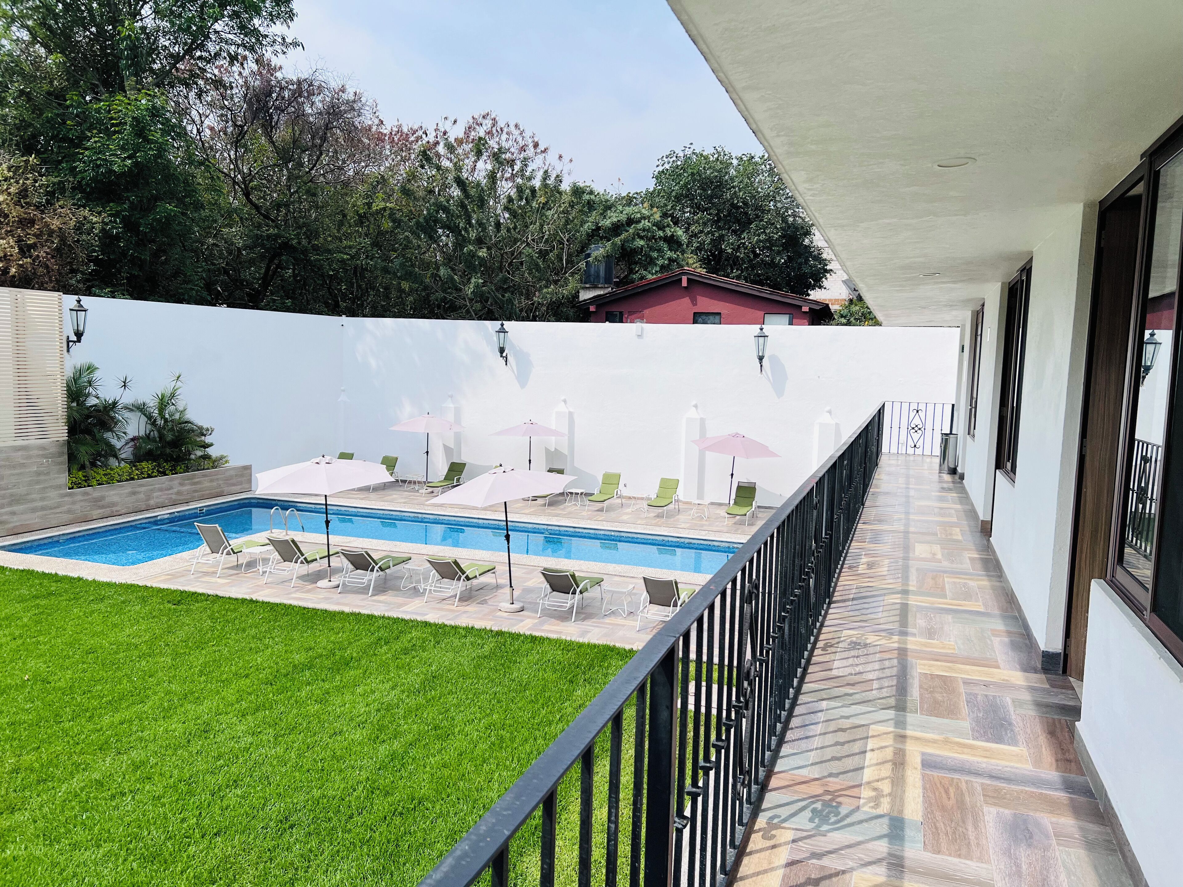 Piscina com espreguiçadeiras e guarda-sóis, rodeada por um muro branco e um espaço gramado.