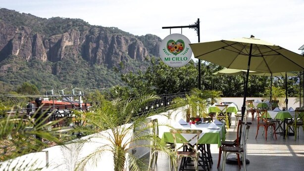 Un área de comedor al aire libre con mesas y sillas, un letrero que dice 'MI CIELO Restaurant', y montañas al fondo.