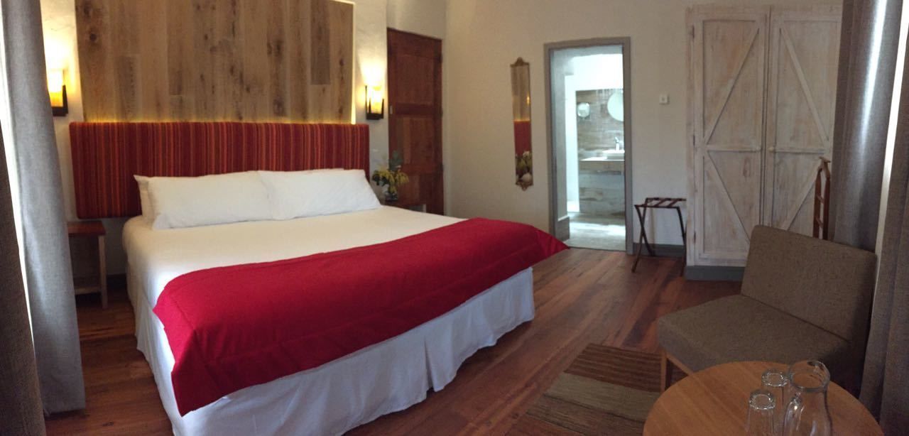 Une chambre d’hôtel avec un grand lit, une tête de lit en bois, un plaid rouge, un canapé et une petite table avec des verres.