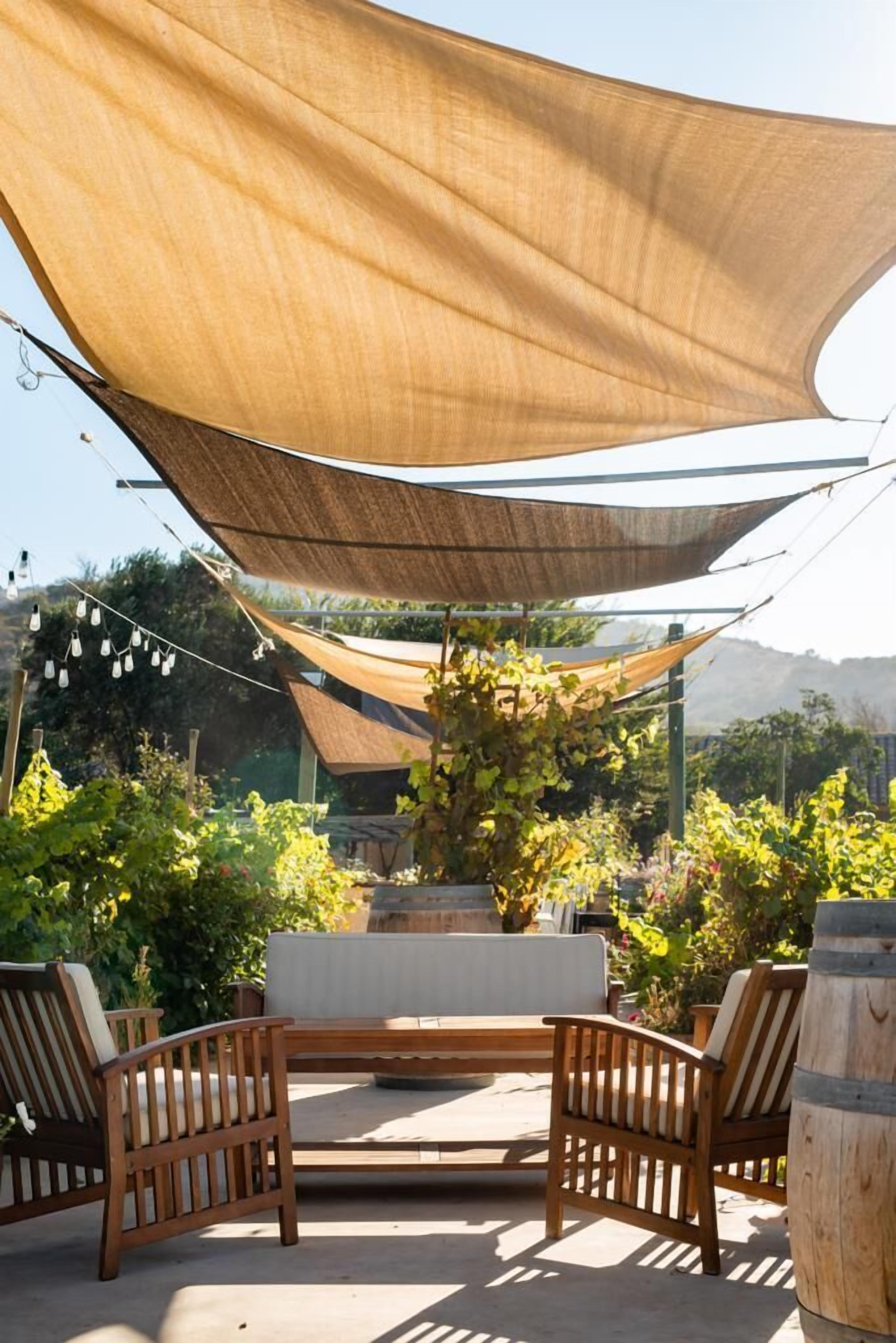 Un área de estar al aire libre con sillas de madera y un banco, protegida por un dosel grande color arena.