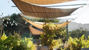 Un área de estar al aire libre con sillas de madera y un banco, protegida por un dosel grande color arena.