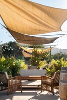 Un área de estar al aire libre con sillas de madera y un banco, protegida por un dosel grande color arena.